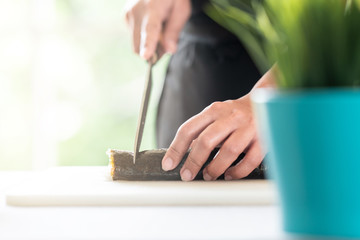 Chef preparing sushi. Asian woman chef in black uniform, about to slice finished roll into pieces with plants in foreground and copy space.