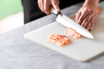 Chef slicing raw salmon on plastic plate. Asian woman chef in black uniform, slicing small piece of salmon. Complete pieces on side with green floor.
