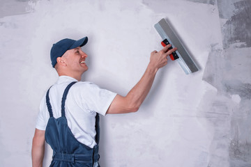 Smiling worker in profile wearing overalls and cap plastering a wall with finishing putty using a...