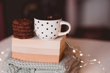 Tasty chocolate cookies with fresh hot tea on open books and knitted sweater closeup over Christmas lights in room. Good morning. Breakfast. School time.