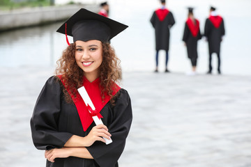 Happy African-American student in bachelor robe and with diploma outdoors