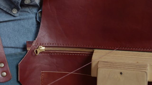 Close up of hands stitching leather piece and zipper with thread and pair of needles