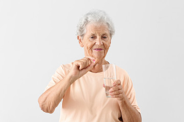 Elderly woman with pill and glass of water on light background
