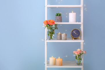 Burning candles with rose flowers on rack near light wall