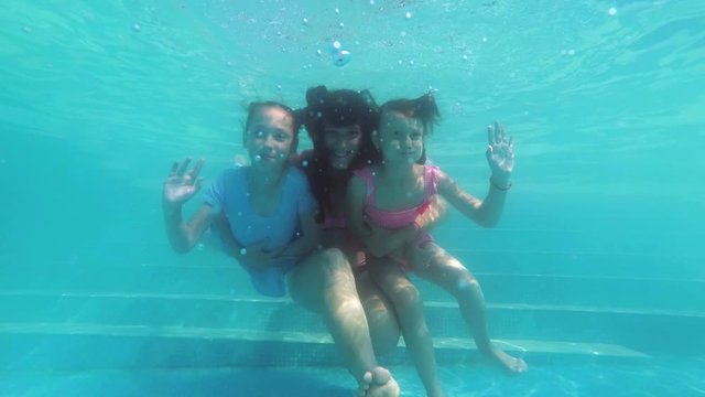 Family: a mother and two young daughters swim and pose underwater at the bottom of the pool on a Sunny day. They hug, look at me and smile. Children waving their hands. Slow motion. Close-up. 4K.