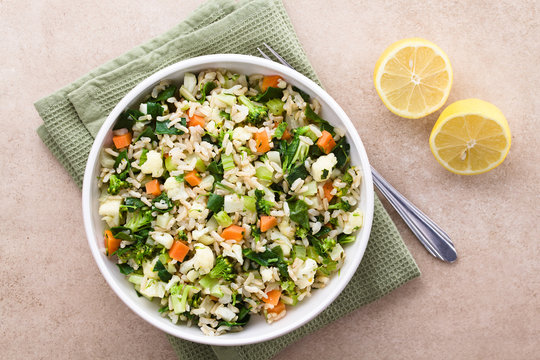 Fresh cooked brown rice with steamed vegetables (broccoli, cauliflower, swiss chard, carrot, celery) in bowl, photographed overhead with lemon on the side