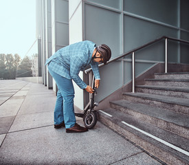 Modern trendy man is locking his electrical scooter near his job at street.