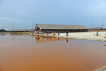 Far shot of workers carrying sea salt baskets and walking on the ramp from sea salt field to barn with reflection on water.