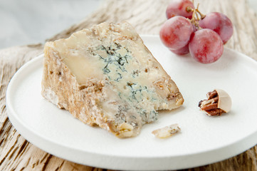 A slice of blue aged Stilton cheese on a wooden table. Cheese is served with large grapes. Quality of agricultural products of farmers. Delicious English cheeses from Russian farmers