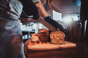 Expirienced baker in protective gloves is slicing bread for daily breakfast at restaurant.