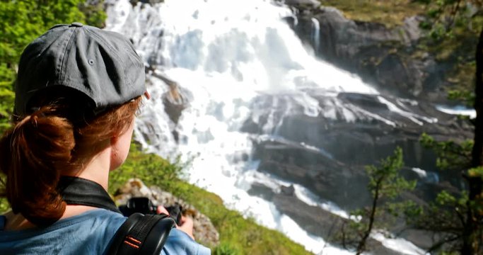 Kinsarvik, Hordaland, Norway. Young Woman Lady Tourist Traveler Taking Pictures Of Waterfall Nyastolfossen In Hardangervidda Mountain Plateau. Nyastolsfossen Is Famous Popular Destination