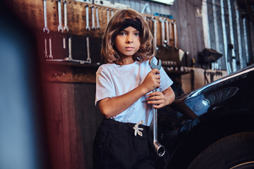 Fototapeta na wymiar Portrait of little cheerful girl with big wrench in hands near shiny gar at auto service.