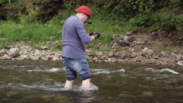 Man takes photos of river water. Stock footage. Professional photographer takes pictures of river flow for travel magazine. Man stands knee-deep in water and taking pictures of nature