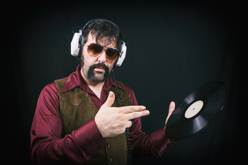 Man in vintage 1970's era clothing, including a vest, sunglasses and mustache. Holding and pointing...