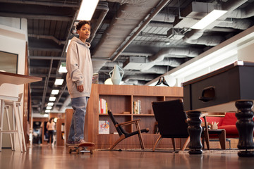 Fototapeta na wymiar Portrait Of Casually Dressed Young Businesswoman With Skateboard Standing In Open Plan Workplace