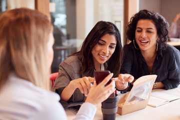 Group Of Young Businesswomen Sitting Around Table Having Working Lunch And Looking At Mobile Phones