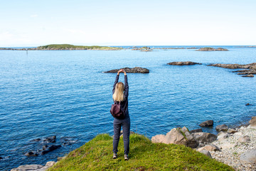 Fototapeta na wymiar Women with backpack looks at the blue ocean in Norway. Enjoys scenic view landscape. Beautiful nature. Harmony, relax lifestyle. Travel, adventure. Sense of freedom. Explore North Norway. Scandinavia