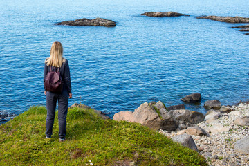 Fototapeta premium Women with backpack looks at the blue ocean in Norway. Enjoys scenic view landscape. Beautiful nature. Harmony, relax lifestyle. Travel, adventure. Sense of freedom. Explore North Norway. Scandinavia