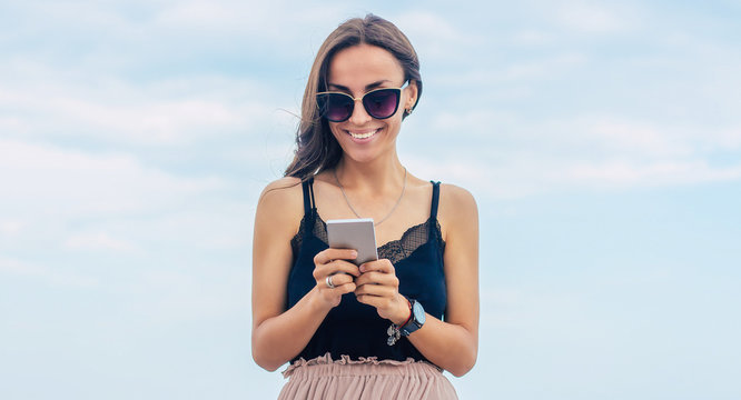 Vacation, travel, relaxation, freedom and happy time. Beautiful smiling young brunette woman in sunglasses is using her smart phone on the sea background