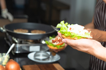 Cook preparing burger adding sauce, vegetable salad, onion and tomato on bread with cream cheese in modern kitchen.