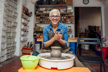 Senior female potter working on pottery wheel while sitting  in her workshop - obrazy, fototapety, plakaty