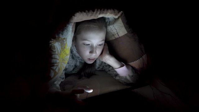 A little girl playing on a tablet at night on a bed under a blanket. Concept video. Close-up. Raw video. 4K.