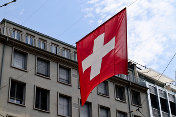 Fototapeta na wymiar Flag of Switzerland hanging on middle of the street with building and blue cloudy sky in background 