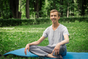 Obraz premium Young attractive man in sport clothes is meditating in the lotus position with a pacified face in the park against the background of green grass and trees.