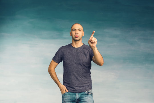 A young bald man in a gray T-shirt on a blue background surprised looking at the camera, raising his index, as if he wants to click on an invisible screen.