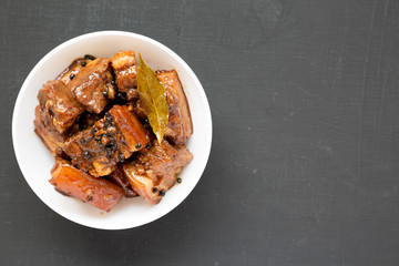 Homemade Filipino Adobo Pork in a white bowl on a black background, top view. Flat lay, overhead, from above. Copy space.