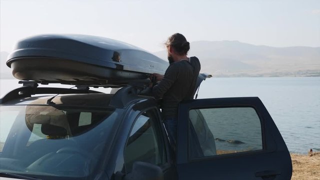 Young bearded man is opening roof bag on his crossover and taking out a camping equipment. Traveler is came to the lake and preparing for camping - gets a tent and from car roof box