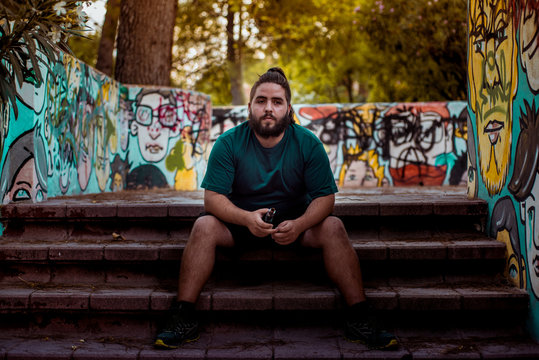 Young man with long hair and big beard sitting on the stairs of a park with graffiti.