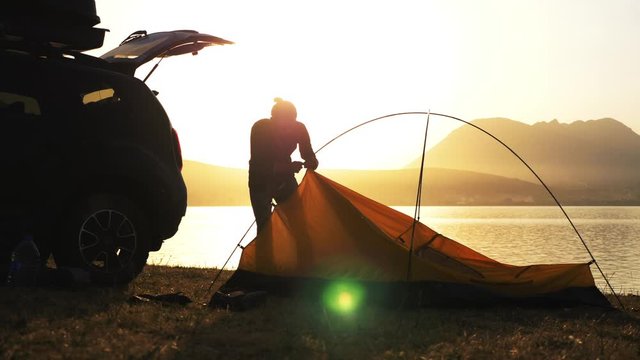 Silhouette of young bearded man sets up a tent at campsite on the lake shore. Traveler is came to the on his crossover and getting ready for the night