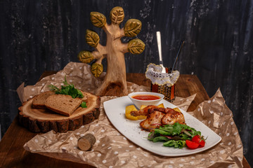 chicken with potatoes, tomatoes and herbs, a Cup of sauce on a white plate and pieces of black bread on a tray of a bar of wood