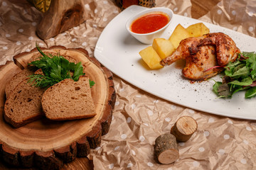 chicken with potatoes, tomatoes and herbs, a Cup of sauce on a white plate and pieces of black bread on a tray of a bar of wood