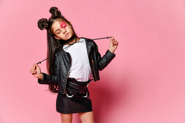 Little hipster girl in a black leather jacket posing on a pink background, holding the ties from the necklace.