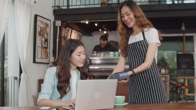 Young Asian freelance women pay contactless credit card at coffee shop. Asian happy girl barista waiter wear gray apon holding credit card reader machine for customer can pay with technology in cafe.