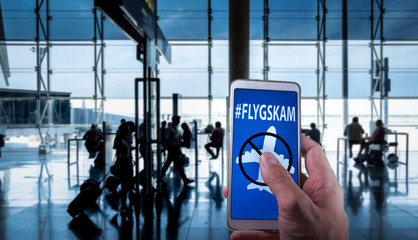 Hand holding a smartphone with Flyskam message on screen with airport passengers on the background....