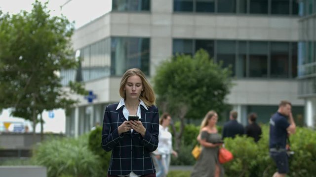 In a clean urban setting, a young women walks towards the camera paying extreme attention to her cell phone