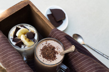 Two glass cups with banana, chocolate, yogurt and smoothie in a wooden box