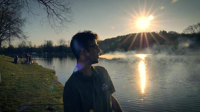 Young man with glasses standing beside a lake at sunset smoking a cigarette exhales blowing a stream of smoke from his mouth.