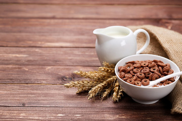 Chocolate corn flakes in bowl with milk and wheat ears on wooden table