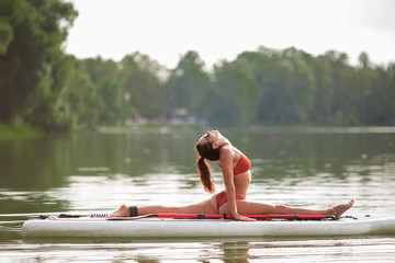 Young pretty fitness girl in swimwear is doing yoga on the SUP in the middle of the lake with a green forest in the background.