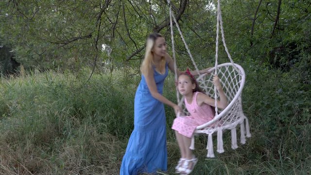 Two young sisters play and swing on a suspended swing in the park, outdoors on a sunny day. They smile and pose in colorful dresses. Fashion portrait. Close-up. 4K.