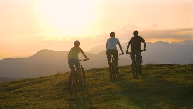 Three friends ride their electric bicycles up to the edge of a grassy hill and high five at sunset. Mountain bikers celebrate at sunset after a successful mountain biking journey in scenic Slovenia.