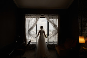 Beautiful young woman in a wedding dress, standing back, and revealing curtains in dark room and looking through the window at the city panorama. Silhouette of young bride posing near the window.