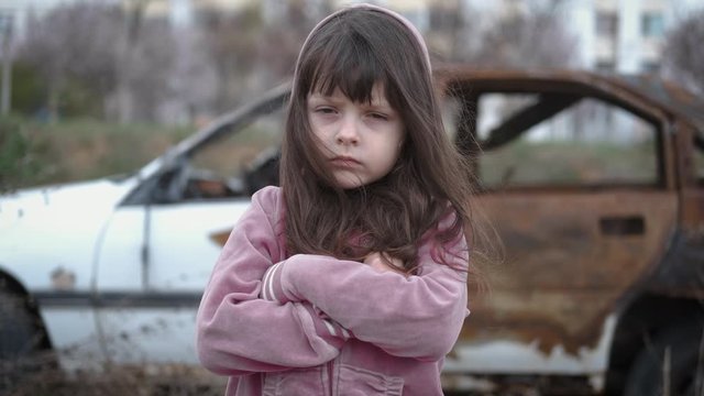 Sadness orphan. Sad little girl on the background of a burnt car. Homeless childhood concept. Depression of an orphan child.