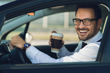 Smiling businessman driving a car while drinking coffee. He is looking at camera