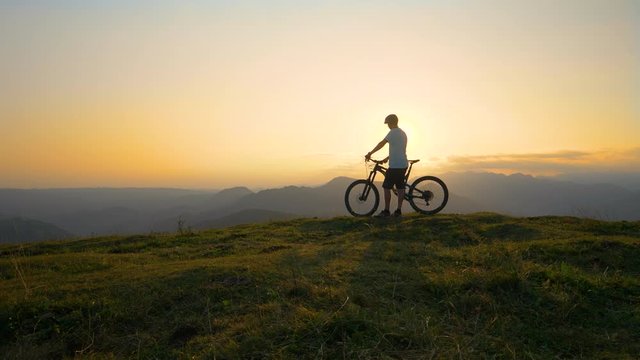 COPY SPACE, AERIAL, SUN FLARE: Golden morning sunbeams shine on the unrecognizable mountain biker standing on top of a grassy hill before downhill riding in the breathtaking Slovenian mountains.