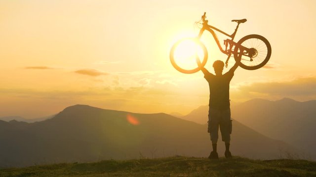 SILHOUETTE SLOW MOTION SUN FLARE COPY SPACE: Golden summer evening sun rays illuminate the bicycle rider celebrating finishing a long cross country trip at sunset. Excited man lifts bicycle above head